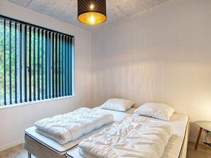 A bedroom with two beds, a window with vertical blinds, a hanging light fixture, and a small table. - 5 Star Holiday Home in Vaeggerlose (Vaeggerlose)