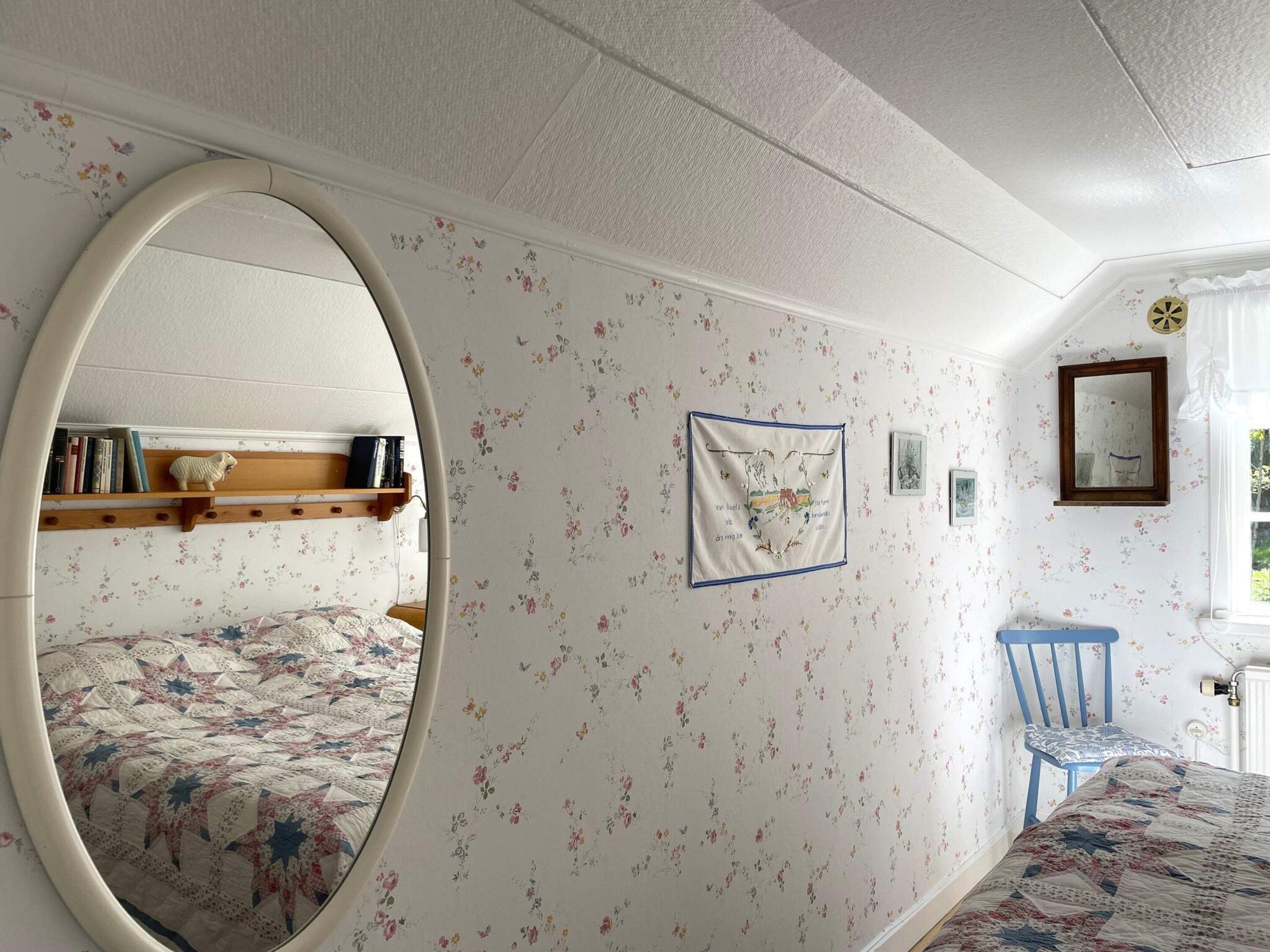 A bedroom with floral wallpaper, a round mirror, a bed with a patterned quilt, a blue chair, and a small shelf with books and a decorative item.