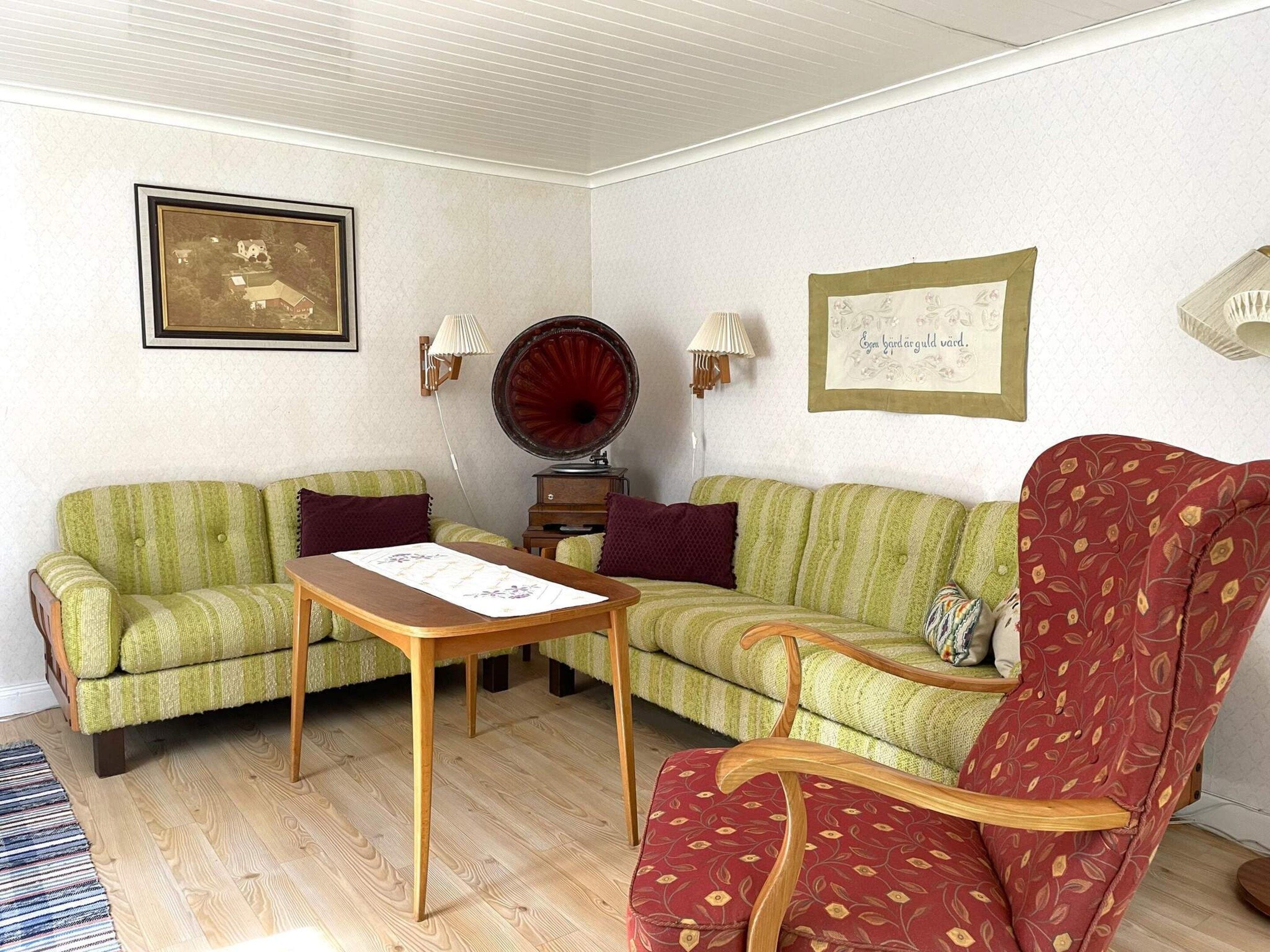 Una sala de estar con dos sofás verdes, una mesa de centro de madera, un sillón rojo estampado, un gramófono y cuadros enmarcados en las paredes.