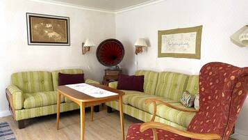 Una sala de estar con dos sofás verdes, una mesa de centro de madera, un sillón rojo estampado, un gramófono y cuadros enmarcados en las paredes.