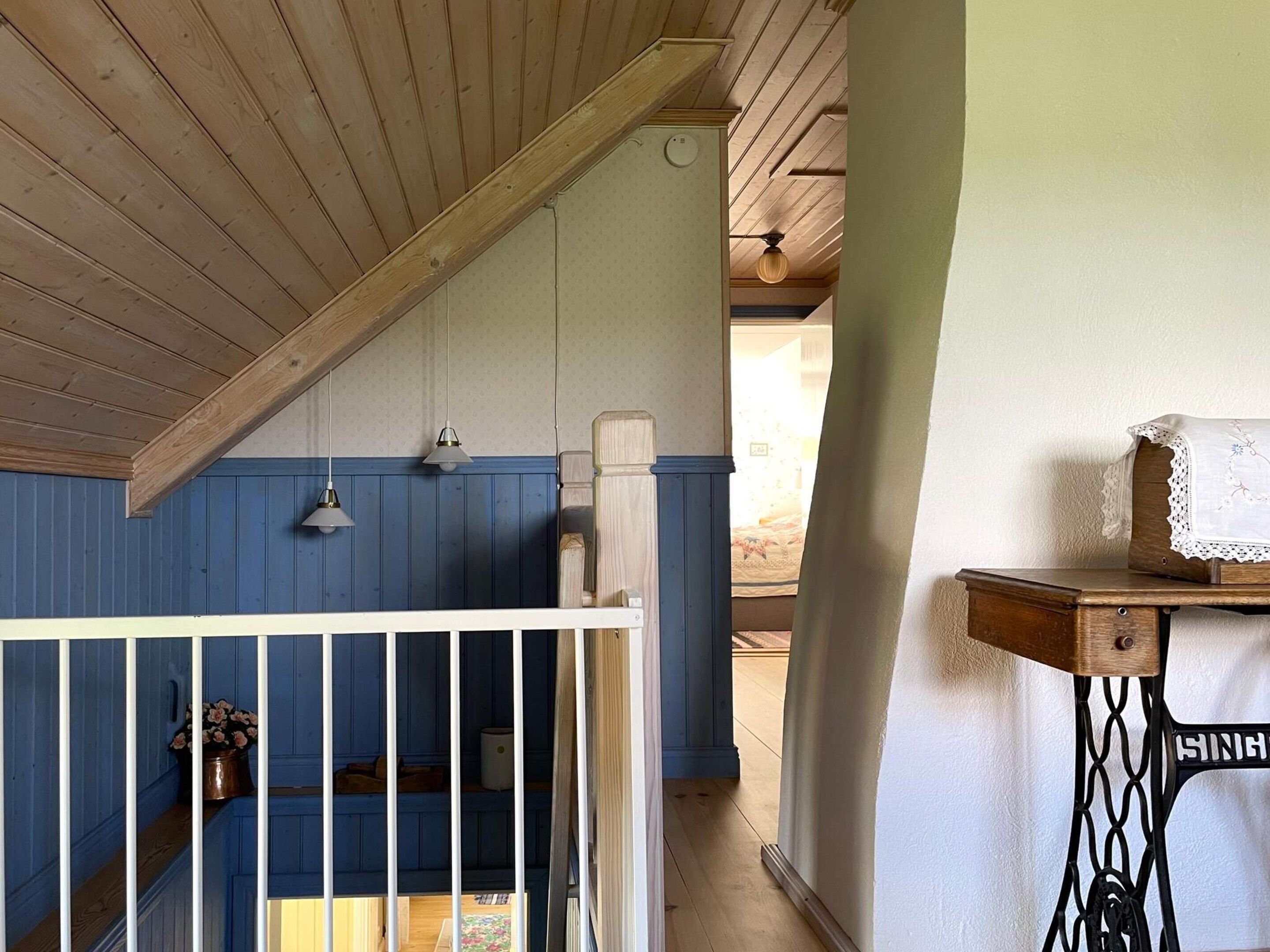 A room with a sloped ceiling, a blue and white wall, a white railing, a sewing machine, and a bed with a floral arrangement.