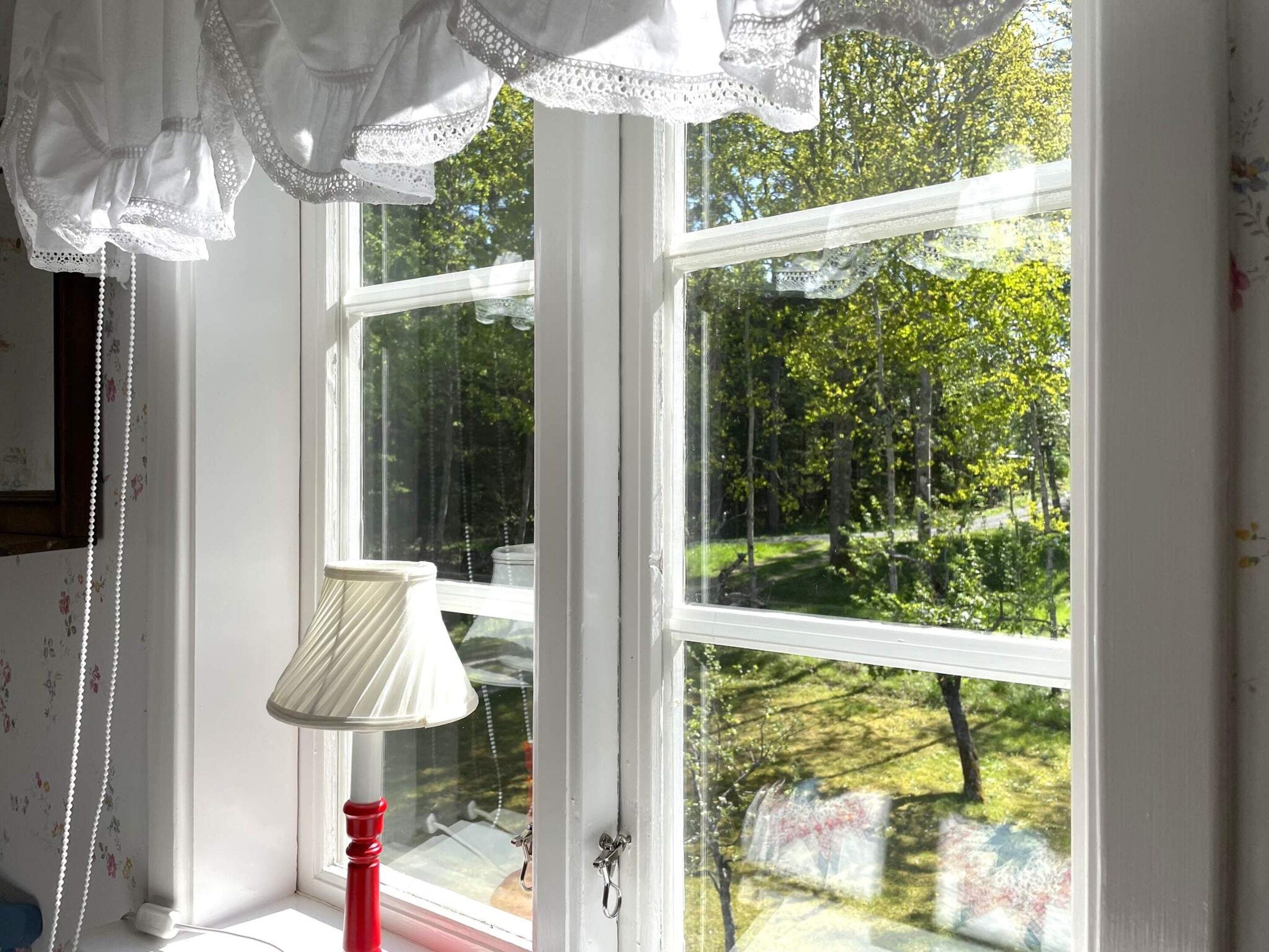 A room with a window overlooking a garden, a white lamp, and a wall with floral wallpaper.