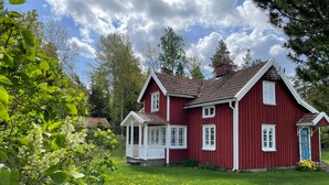 Een rode woning met veranda en een blauwe deur, omringd door groen gras en bomen.