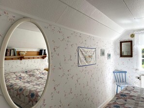 A bedroom with floral wallpaper, a round mirror, a bed with a patterned quilt, a wooden shelf with books, and a blue chair. - 6 Person Holiday Home in Grastorp (Grastorp)