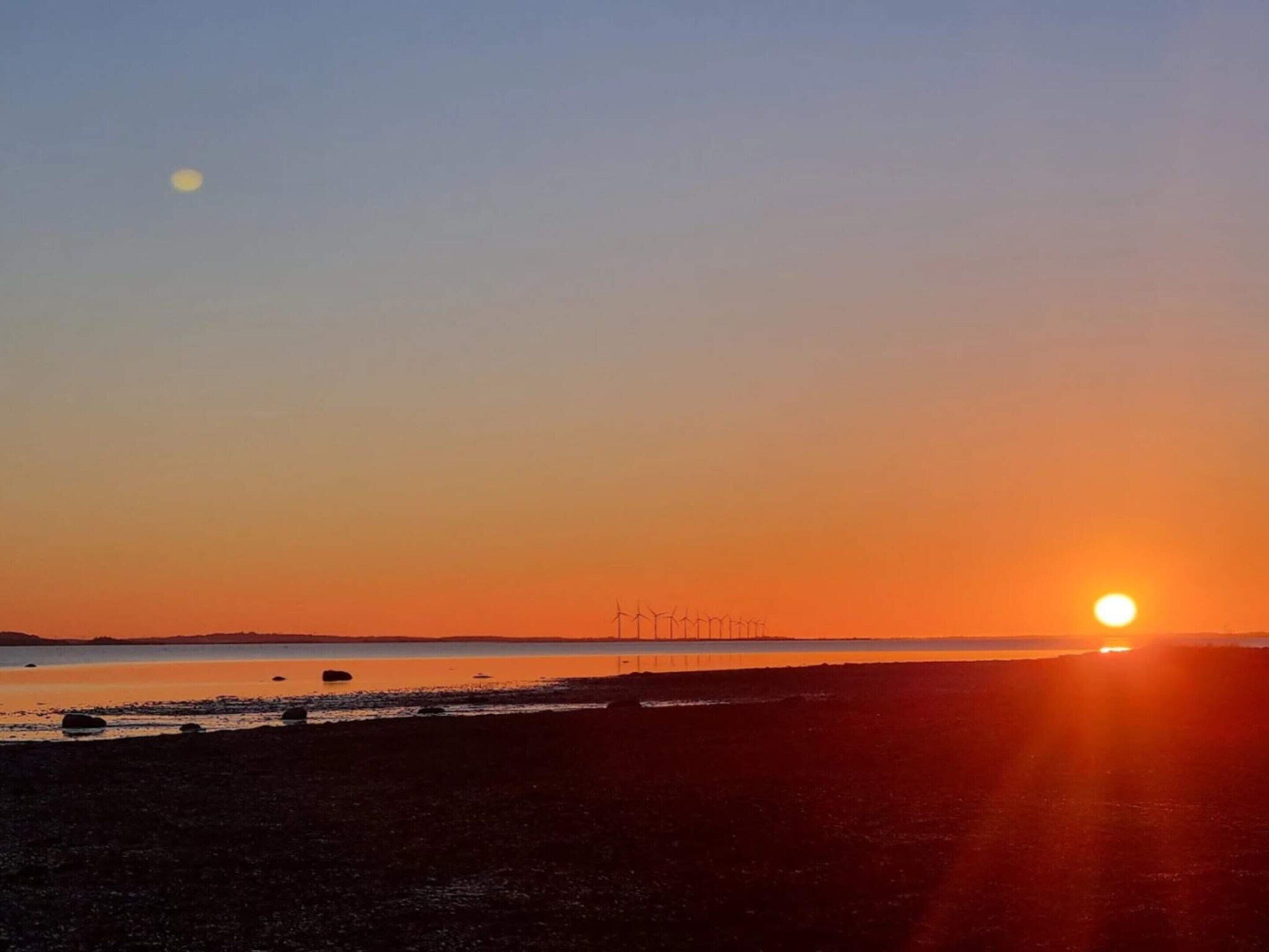 Auringonlasku rannalla, ja kauempana horisontissa näkyy tuulivoimaloita.