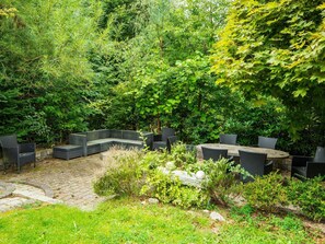 Un espace extérieur aménagé avec une terrasse en pierre, entouré d’une végétation luxuriante et d’arbres.