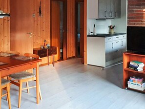 A modern interior with a wooden dining table, chairs, a kitchen area with cabinets and a television, and a bookshelf.