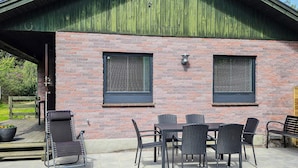 A brick building with a green roof, two windows with blinds, a covered patio with a table and chairs, and a bench. - 6 Person Holiday Home in Hojby (Hojby)