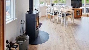 A cozy room with a wood-burning stove, a dining table with chairs, a television, and large windows offering a view of greenery outside. - 7 Person Holiday Home in Hojslev (Hojslev)