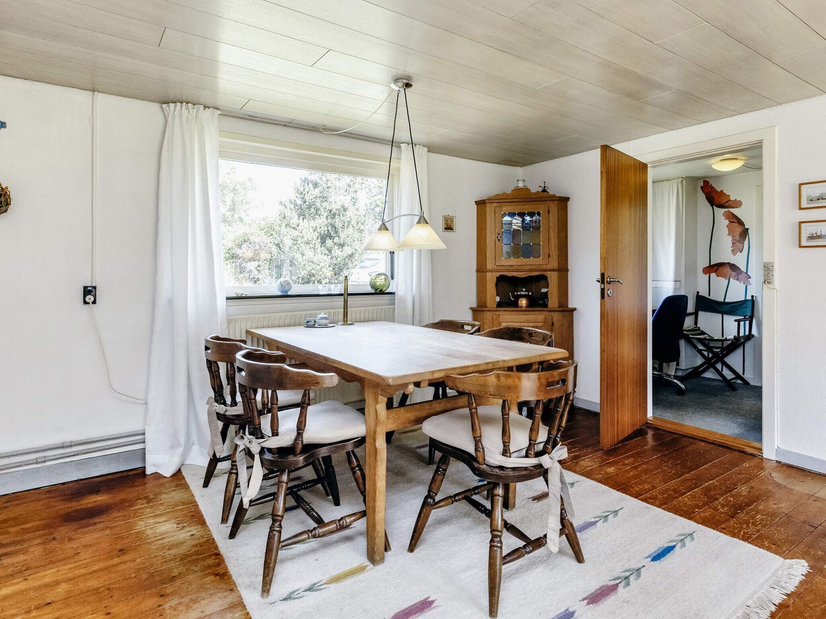 Un coin repas avec une table et des chaises en bois, un radiateur, une fenêtre avec des rideaux, un meuble et une porte donnant sur une autre pièce.