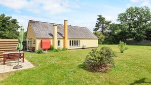 Une maison de plain-pied avec un toit en pente, une cheminée et un petit jardin agrémenté d’un banc et d’une table.