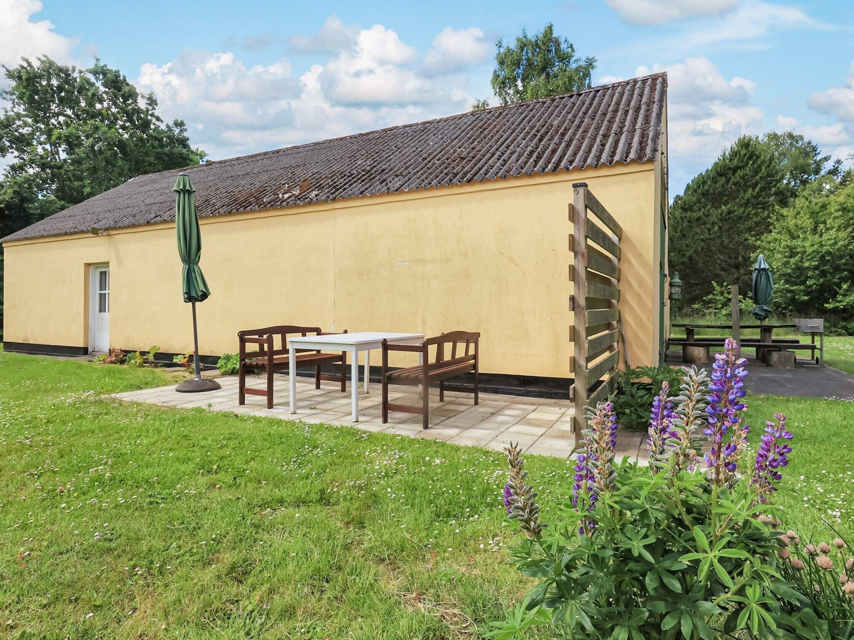 Un petit bâtiment avec un toit en tuiles, une porte et une fenêtre. On y trouve une table et deux chaises à l’extérieur.