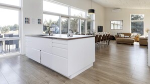 A modern kitchen with large windows, a dining area with a table and chairs, and a living room with a sofa and a coffee table. - 10 Person Holiday Home in Vaeggerlose (Vaeggerlose)