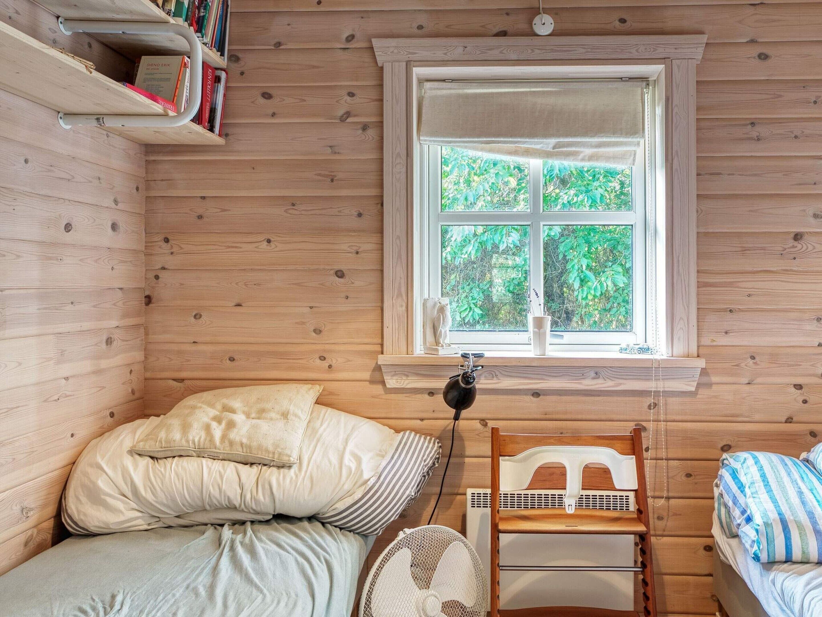 A cozy cabin room with a bed, a chair, a window with a view of greenery, and a shelf with books.