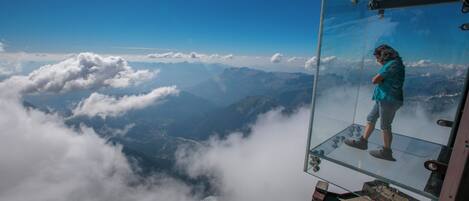 A glass observation platform with a person standing on it, overlooking a mountainous landscape with clouds below.