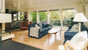A spacious living room with a large sofa, a coffee table, and a dining area with a table and chairs. There is a large window providing a view of the outdoors.