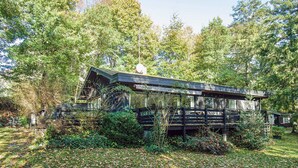 A rustic cabin surrounded by dense forest, with a wooden deck and a satellite dish on the roof.