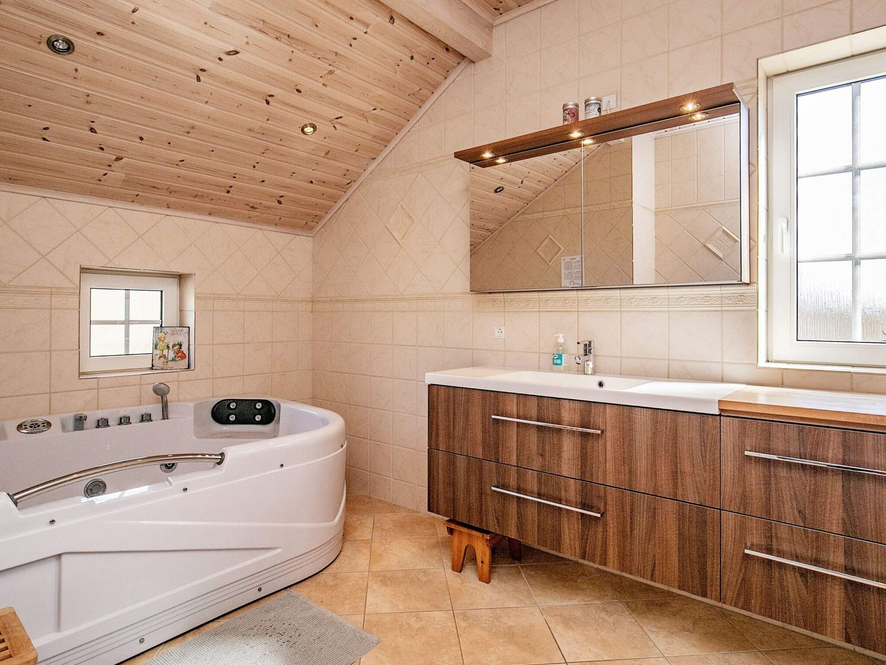 A modern bathroom with a wooden ceiling, a large bathtub, a vanity with drawers, a mirror, and a window.