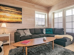 Uma sala de estar aconchegante com um sofá seccional cinza escuro, uma mesa de centro de madeira e um tapete preto e branco. Há duas janelas com persianas, um quadro na parede e um abajur em uma mesa lateral.