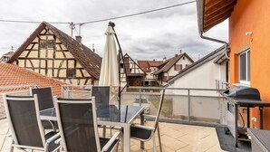 Une terrasse avec des meubles d’extérieur, un barbecue, et des maisons traditionnelles en arrière-plan.