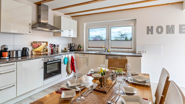 Une cuisine moderne avec des meubles blancs, un coin repas doté d’une table en bois pour deux personnes et une grande fenêtre avec des stores.