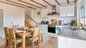 Une cuisine moderne avec des poutres en bois, des armoires blanches, un évier en acier inoxydable et un coin repas avec des chaises et une table en bois, le tout disposé pour accueillir quatre personnes.