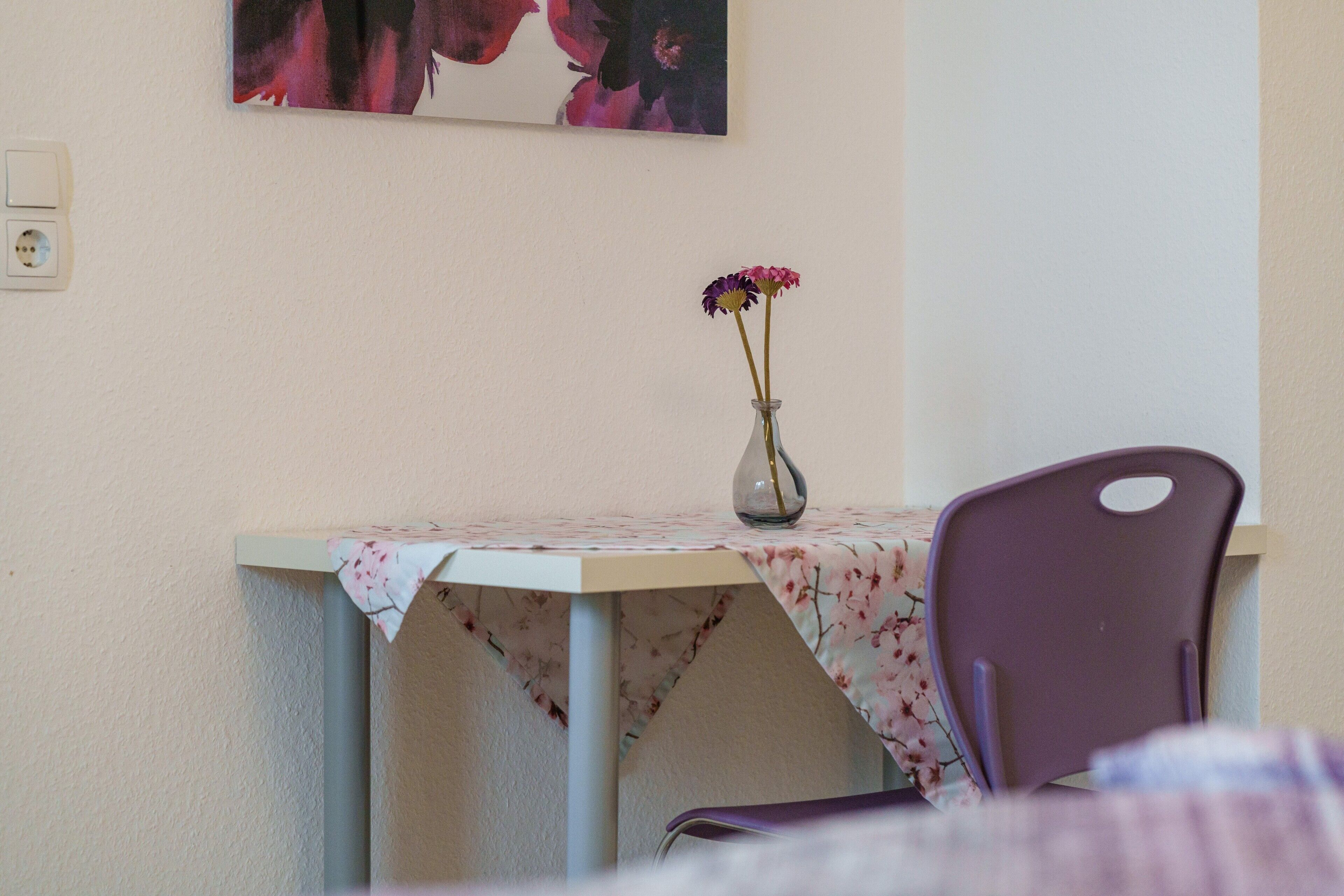 Ein Esstisch mit Blumen in einer Vase, ein Stuhl und ein Wandbild.