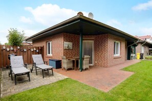 Ein Ziegelhaus mit überdachter Terrasse, zwei Liegestühlen, einem Grill und einem eingezäunten Garten.