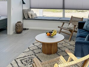 Una sala de estar moderna con una mesa redonda, sillas de mimbre, un sofá con cojines y vistas a través de grandes ventanales.