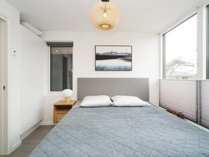 Un dormitorio con cama, mesita de noche con lámpara, ventana y un cuadro colgado en la pared.