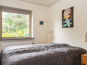 Une chambre à coucher avec un lit, une fenêtre donnant sur un paysage verdoyant, une lampe fixée au mur et un tableau.