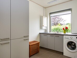 A compact laundry room with a washing machine, a small wooden basket, and a window with a view of greenery. - 8 Person Holiday Home in Struer (Struer)