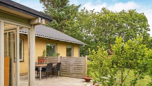 Et hus med en terrasse, et bord og stole, omgivet af grønne planter og træer.