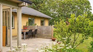 Et hus med en terrasse, et bord og stole, omgivet af grønne planter og træer.