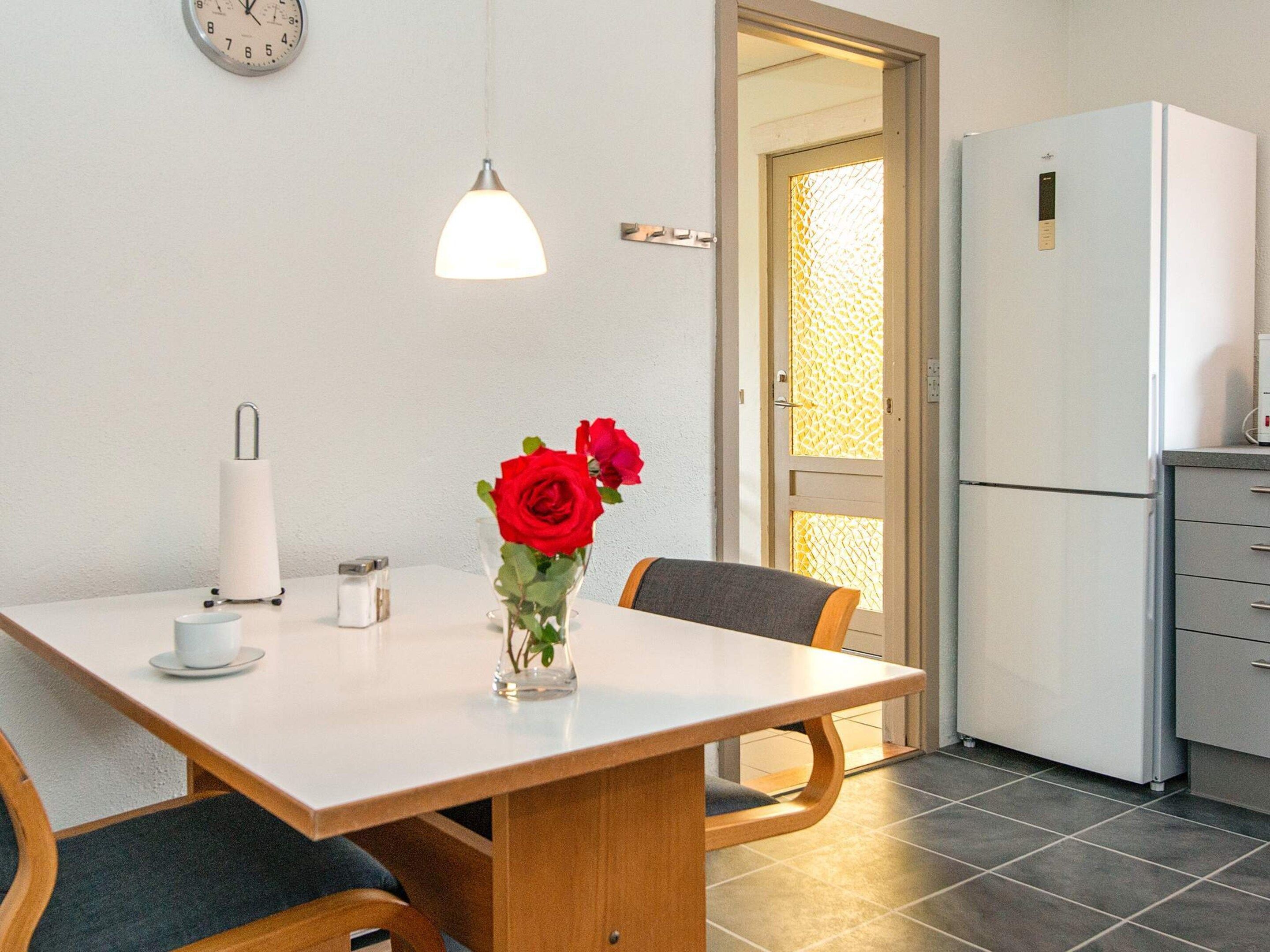 Ein Essbereich mit Tisch, Stühlen, einer Vase mit roten Rosen, einer Uhr und einem Kühlschrank.