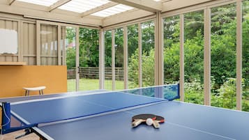 Ein Tischtennis-Tisch mit zwei Schlägern und einem Ball, eine Whiteboard-Tafel und ein Holzschemel in einem Raum mit großen Fenstern und Blick auf einen Garten.