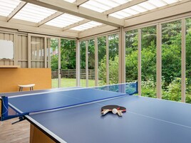 A ping pong table with two paddles and a ball, a whiteboard, and a wooden stool in a room with large windows and a view of a garden.