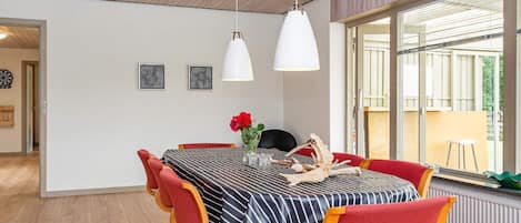 Uma sala de jantar com uma mesa posta para quatro pessoas, cadeiras de madeira, um vaso com uma flor vermelha e uma grande janela com vista para o exterior.