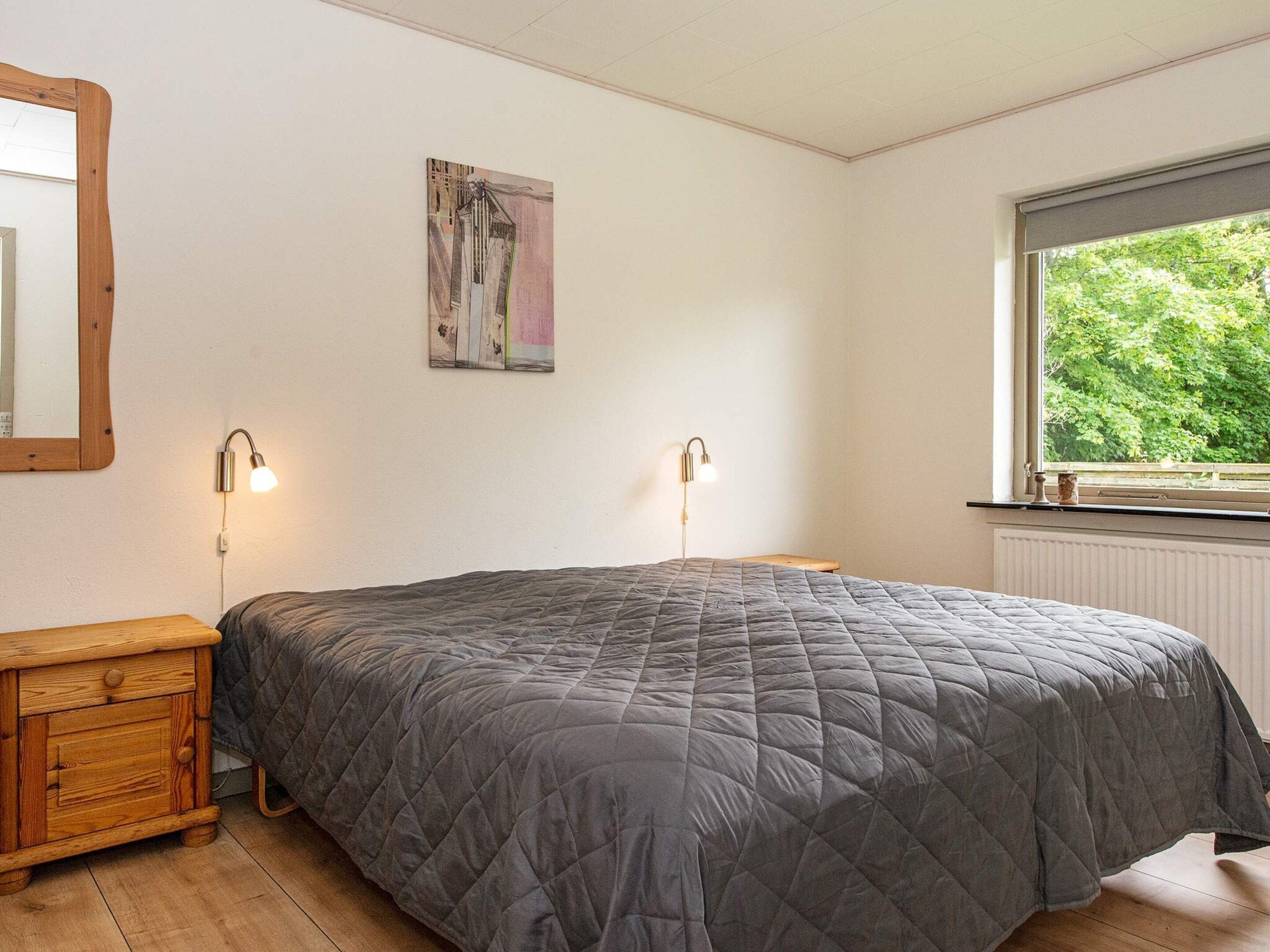 A bedroom with a bed, bedside lamps, a wooden nightstand, a painting on the wall, and a window with a view of trees.