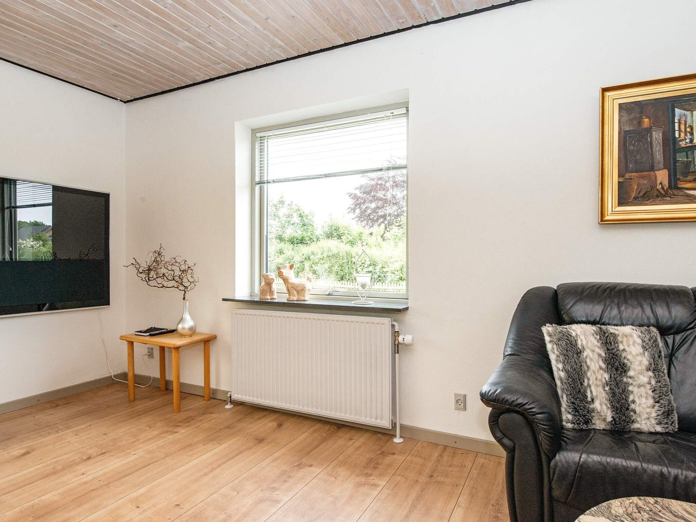 Ein Zimmer mit einer schwarzen Ledersofa, einem Holznachttisch mit einer Vase, einem Heizkörper und einem Fenster mit Blick auf Grünflächen.