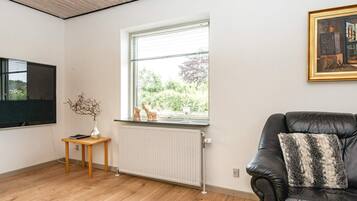 Et værelse med en sort lædersofa, et træsidebord med en vase, en radiator og et vindue med udsigt til grøn vegetation.
