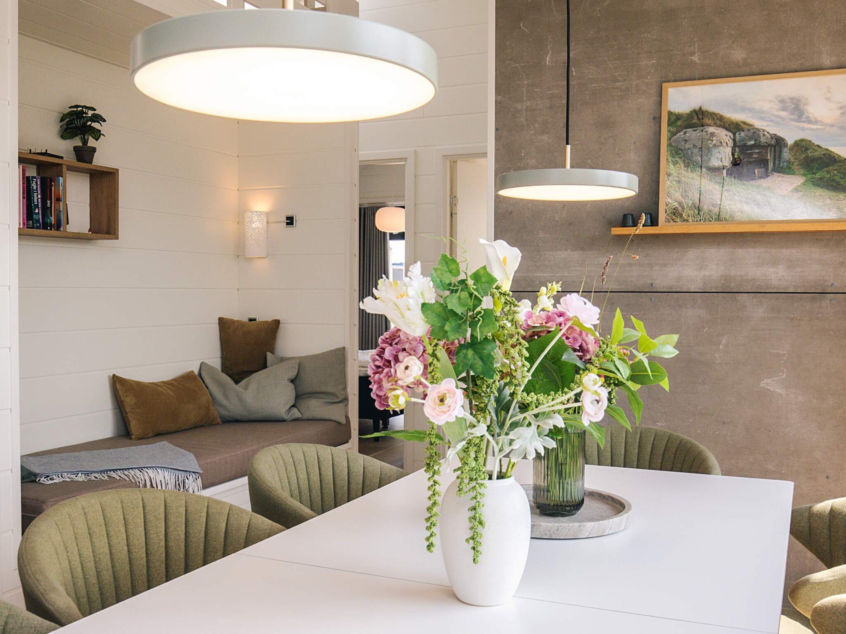 A modern living room with a white sofa, green chairs, a white table with a vase of flowers, and a painting on the wall.