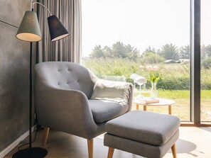Una habitación moderna con un sillón gris y reposapiés, una lámpara de pie y vistas a zonas verdes exteriores.
