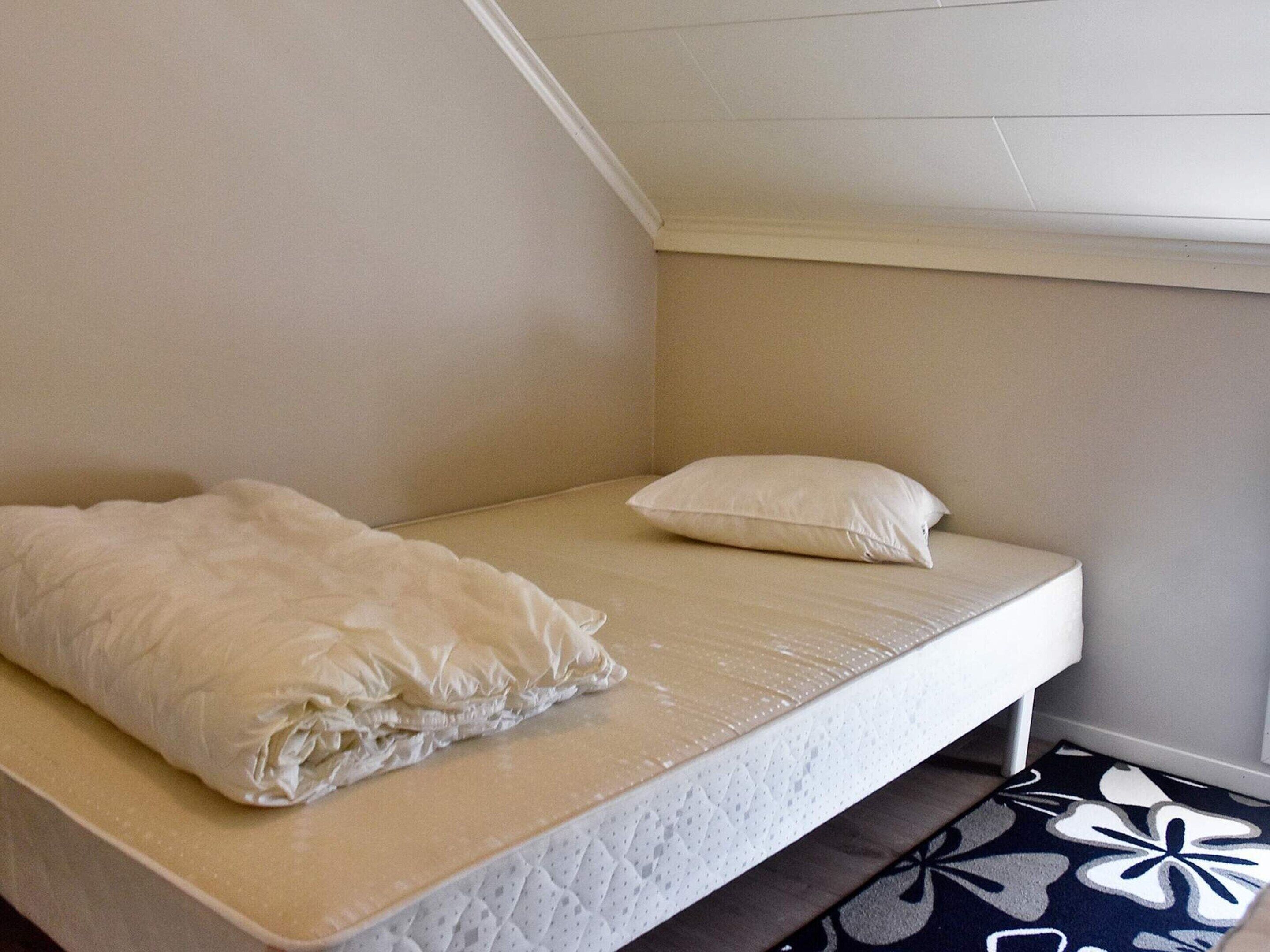 Een enkel bed met een witte matras en kussen, een beige muur en een kamerbreed tapijt met patroon.