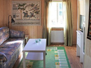 A room with a sofa, a small table, a radiator, a television, and a window with curtains. - 6 Person Holiday Home in Stjarnhov (Stjarnhov)