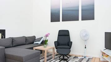 Una sala de estar moderna con un sofá seccional gris, una silla de oficina negra, una mesita con una orquídea en maceta, un ventilador de pie y un televisor sobre un mueble blanco.