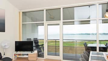Una sala de estar moderna con un televisor de pantalla plana, asientos y un amplio ventanal que da a un balcón con vistas al agua.