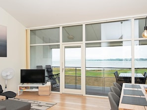 A modern living room with a flat-screen TV, seating, and a large glass door leading to a balcony with a view of a body of water. - 6 Person Holiday Home in Ebeltoft (Ebeltoft)