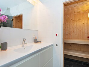 Ein modernes Badezimmer mit weiß gefliester Wand, einem großen Spiegel, einem Waschbecken mit moderner Armatur und einer Holztür zur Sauna.