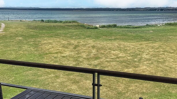A grassy area with a metal railing, a clear view of a body of water, and a distant shoreline.